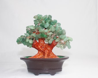 May include: A decorative bonsai tree sculpture. The tree features a reddish-brown trunk and branches, with green stone leaves. It is set in a dark brown, ceramic pot. The tree is a unique home decor item.