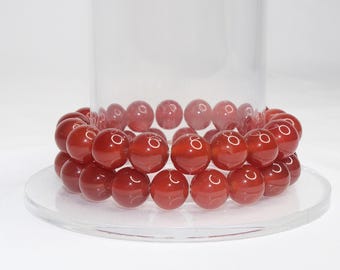May include: A pair of red-orange beaded bracelets. The bracelets are made of large, round beads and are displayed on a clear, round glass plate. The beads have a glossy finish and are arranged in a double-strand design.