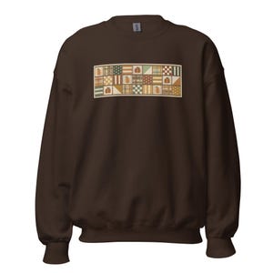 Op de afbeelding: Een donkerbruine sweater met ronde hals en een rechthoekige afbeelding op de borst. De afbeelding bestaat uit een raster van kleine vierkantjes in tinten oranje, groen, crème en bruin, met diverse patronen zoals strepen, stippen en pompoenen.