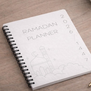 May include: A white spiral-bound Ramadan Planner with the text "RAMADAN PLANNER" and the year "2026" and "1447" printed on the cover. The cover features a line drawing of a mosque with clouds and stars.