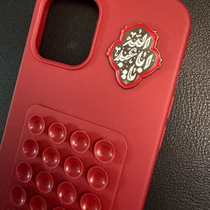 May include: A crimson phone case featuring a textured area of raised red circles. An emblem with Arabic script in black and white is attached. The case is designed to protect a smartphone.