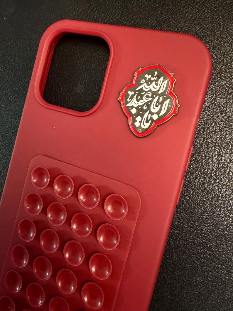 May include: A crimson phone case featuring a textured area of raised red circles. An emblem with Arabic script in black and white is attached. The case is designed to protect a smartphone.