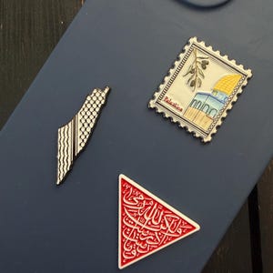 May include: Three enamel pins on a blue phone case. One pin is a map of Palestine with a black and white pattern. Another is a stamp with a building and olive branches. The third is a red triangle with white Arabic script.