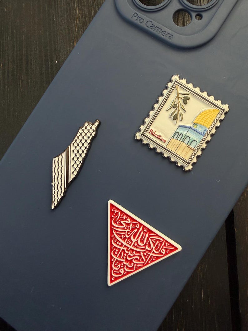 May include: Three enamel pins on a blue phone case. One pin is a map of Palestine with a black and white pattern. Another is a stamp with a building and olive branches. The third is a red triangle with white Arabic script.