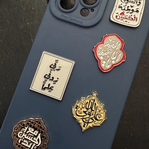 May include: A selection of decorative pins with Arabic calligraphy on a blue phone case. The pins are in various shapes and colours, including gold, silver, and red accents. The phone case has a Pro Camera label.