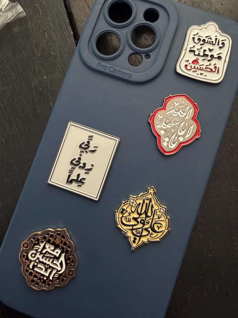 May include: A selection of decorative pins with Arabic calligraphy on a blue phone case. The pins are in various shapes and colours, including gold, silver, and red accents. The phone case has a Pro Camera label.