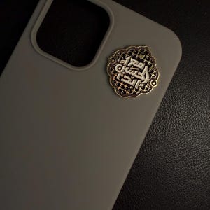 May include: A grey phone case with a decorative gold and silver emblem. The emblem features Arabic calligraphy within an ornate, patterned border. The case has a smooth finish and a camera cutout.