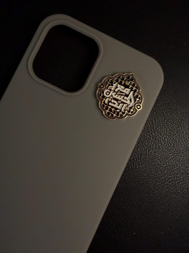 May include: A grey phone case with a decorative gold and silver emblem. The emblem features Arabic calligraphy within an ornate, patterned border. The case has a smooth finish and a camera cutout.
