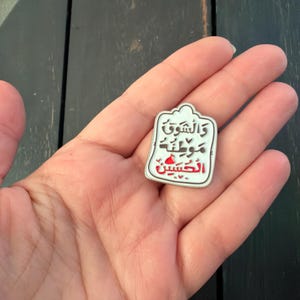 May include: A white and red enamel pin featuring Arabic script. The pin has a distinctive shape with a rounded top and a rectangular body. The text is in black and red. The pin is held in a hand.