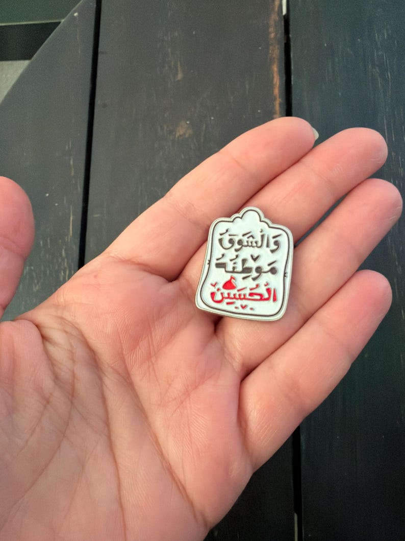May include: A white and red enamel pin featuring Arabic script. The pin has a distinctive shape with a rounded top and a rectangular body. The text is in black and red. The pin is held in a hand.