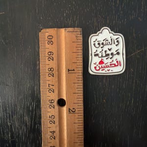 May include: A white and red enamel pin with Arabic text, next to a wooden ruler. The pin has a decorative border and features the words "والشوق موطنه الحسين" in black and red. The ruler is marked in inches, approximately 2.54 cm.