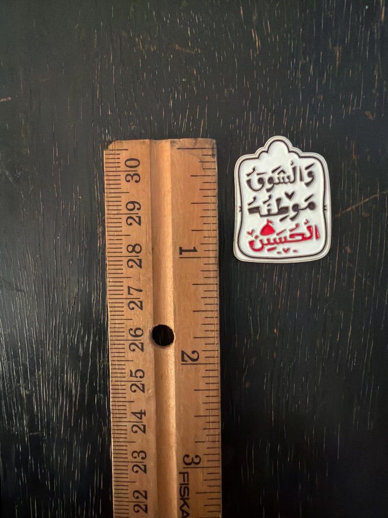 May include: A white and red enamel pin with Arabic text, next to a wooden ruler. The pin has a decorative border and features the words "والشوق موطنه الحسين" in black and red. The ruler is marked in inches, approximately 2.54 cm.