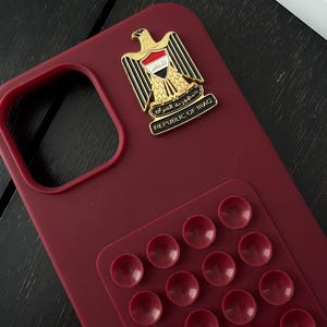 May include: A burgundy phone case with a Republic of Iraq emblem and a pop-up grip. The emblem features a golden eagle with the Iraqi flag and the text "REPUBLIC OF IRAQ". The pop-up grip has multiple burgundy suction cups.
