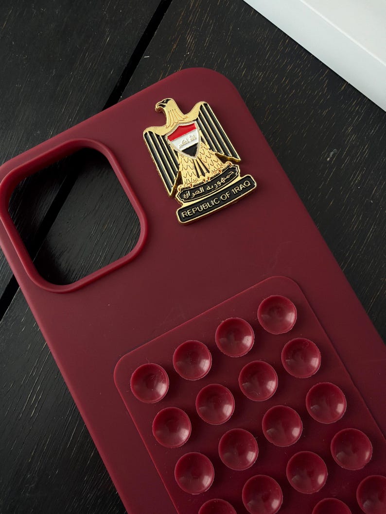 May include: A burgundy phone case with a Republic of Iraq emblem and a pop-up grip. The emblem features a golden eagle with the Iraqi flag and the text "REPUBLIC OF IRAQ". The pop-up grip has multiple burgundy suction cups.