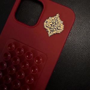 May include: A crimson phone case with a gold emblem bearing Arabic script. The case has a textured finish and a clear section with numerous raised, circular bumps. The emblem is positioned in the top right corner.