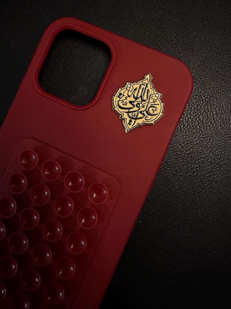 May include: A crimson phone case with a gold emblem bearing Arabic script. The case has a textured finish and a clear section with numerous raised, circular bumps. The emblem is positioned in the top right corner.