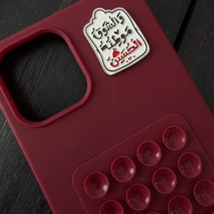 May include: A claret-coloured phone case with a white and red enamel pin attached. The pin features Arabic script. A clear, textured grip pad with multiple suction cups is adhered to the case.