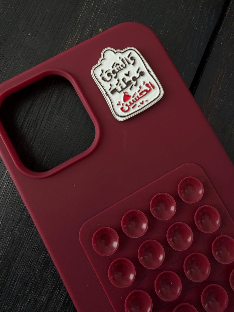 May include: A claret-coloured phone case with a white and red enamel pin attached. The pin features Arabic script. A clear, textured grip pad with multiple suction cups is adhered to the case.