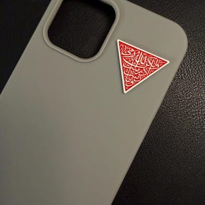 May include: A grey phone case with a red and white triangular emblem. The emblem features Arabic calligraphy. The case has a camera cut-out and is made of a smooth material. The background is dark.