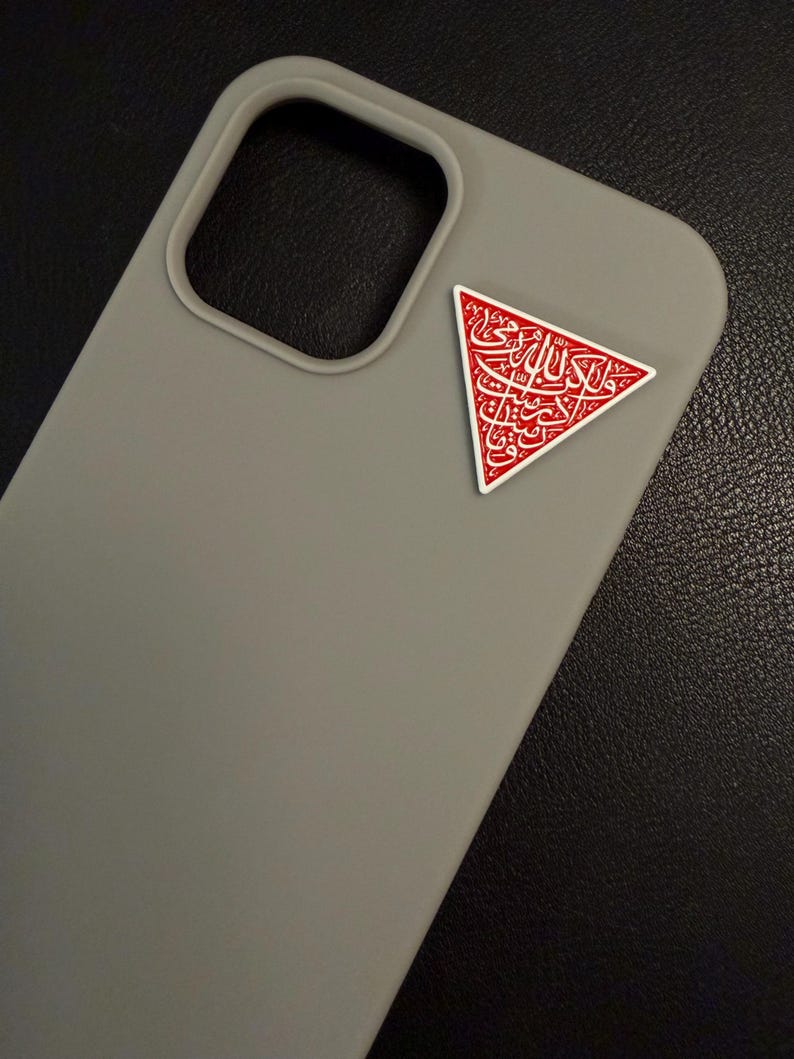 May include: A grey phone case with a red and white triangular emblem. The emblem features Arabic calligraphy. The case has a camera cut-out and is made of a smooth material. The background is dark.