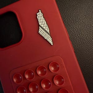 May include: A red phone case with a Palestine map pin. The pin is white and black with a detailed design. A red suction pad with multiple suction cups is attached to the case.