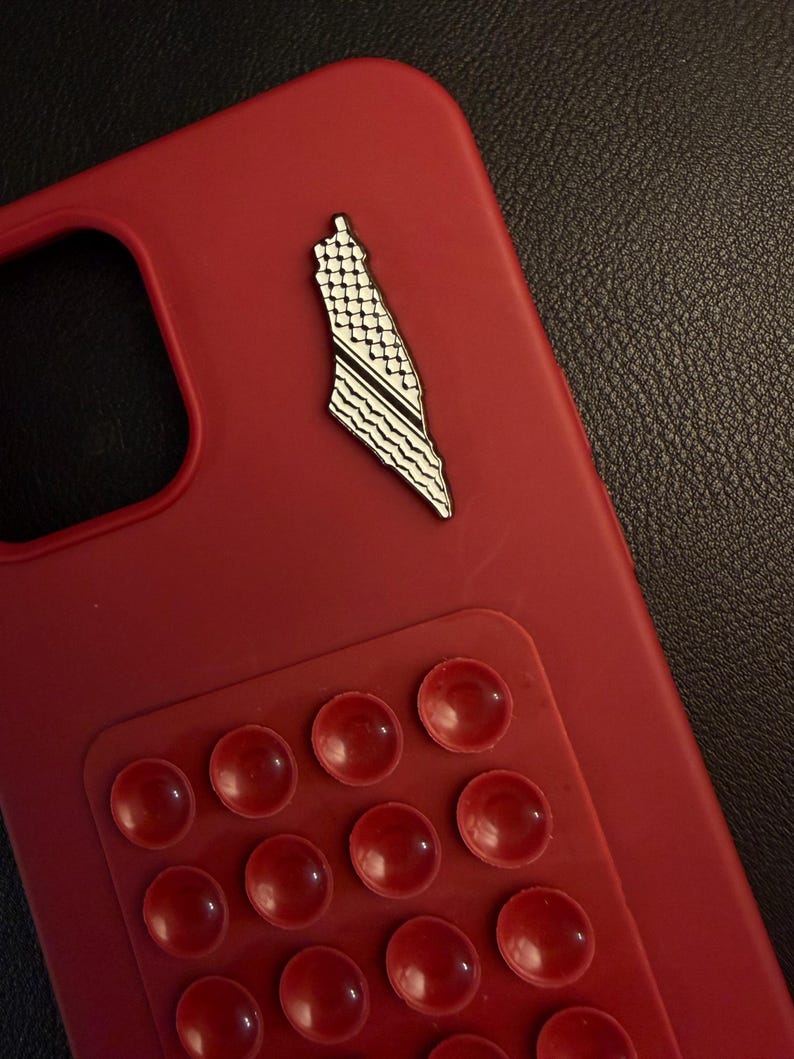 May include: A red phone case with a Palestine map pin. The pin is white and black with a detailed design. A red suction pad with multiple suction cups is attached to the case.