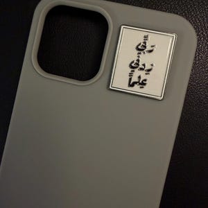 May include: A grey phone case with a rectangular silver emblem. The emblem features black Arabic script. The phone case has a camera cutout and is set against a dark background.