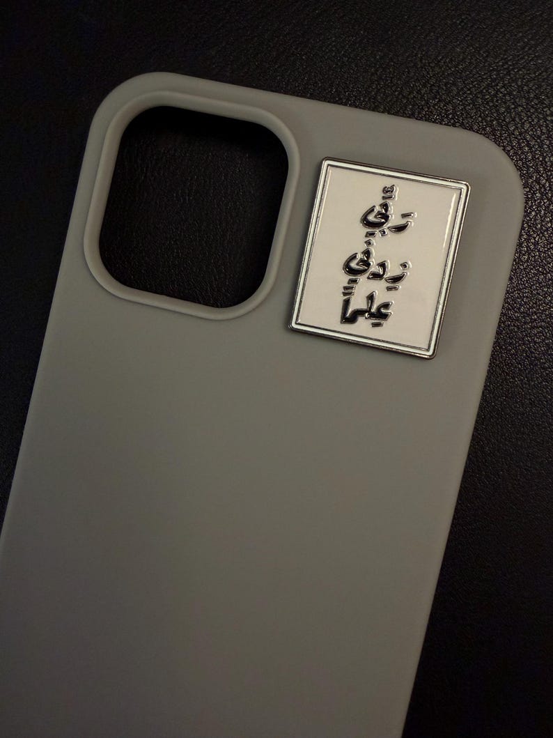 May include: A grey phone case with a rectangular silver emblem. The emblem features black Arabic script. The phone case has a camera cutout and is set against a dark background.