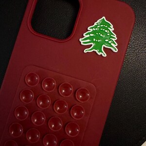 May include: A claret-coloured phone case featuring a green and white cedar tree emblem. The case has a raised, textured section with multiple red, rounded bumps. The case is designed to protect the phone.