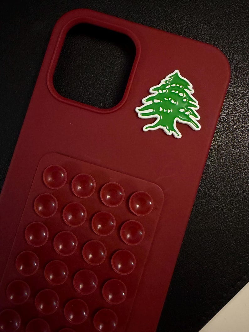 May include: A claret-coloured phone case featuring a green and white cedar tree emblem. The case has a raised, textured section with multiple red, rounded bumps. The case is designed to protect the phone.