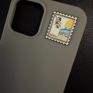 May include: A grey phone case with a decorative stamp showing a yellow-domed building, olive branch, and the word "Palestine". The stamp is attached to the top right of the case.