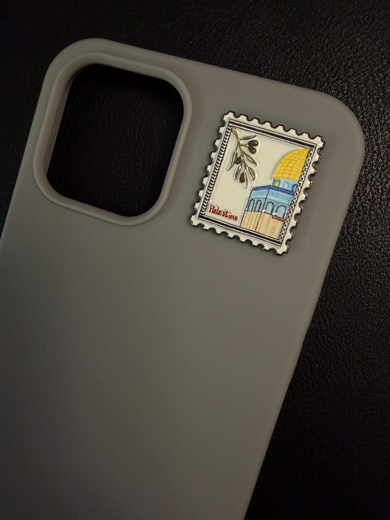May include: A grey phone case with a decorative stamp showing a yellow-domed building, olive branch, and the word "Palestine". The stamp is attached to the top right of the case.