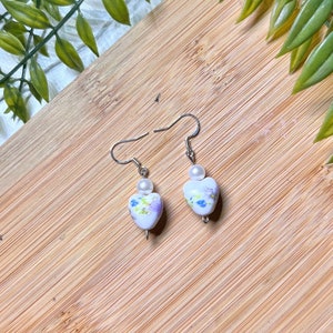 May include: A pair of silver heart-shaped earrings with a white floral design and a single white pearl bead on each earring.