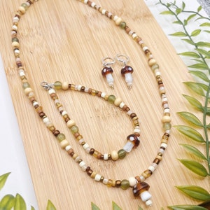 May include: A set of mushroom-themed jewellery featuring a necklace, bracelet, and earrings. The necklace and bracelet are made with brown, white, and green beads. The earrings are silver with brown and white mushroom charms.