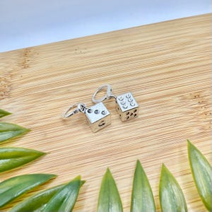 May include: Silver dice earrings. Each earring has a small, cube-shaped die with black dots. The dice are attached to a small hoop. The earrings are shown on a wooden surface with green leaves. The dice are a classic game accessory.