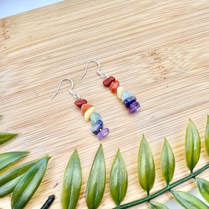 May include: A pair of dangle earrings featuring a chakra design. Each earring has a silver hook and is adorned with a series of colorful, polished gemstones in shades of red, orange, yellow, green, blue, indigo, and purple. The earrings are displayed on a wooden surface.