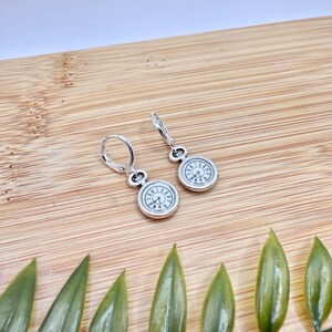 Peut inclure: Boucles d'oreilles argentées avec des cadrans de montre miniatures, des chiffres romains et des aiguilles noirs. Les boucles d'oreilles ont une fermeture à levier et sont présentées sur une surface en bois avec des feuilles vertes.