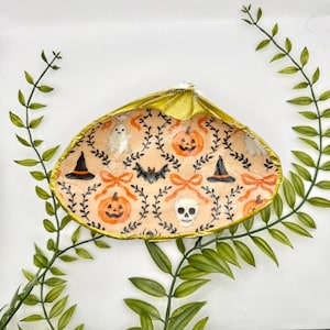 Może przedstawiać: Ozdobna muszla z pozłacaną krawędzią, z motywem Halloween. Wzór zawiera dynie, duchy, nietoperze, kapelusze czarownic i czaszkę, wszystko na brzoskwiniowym tle z pomarańczowymi kokardkami i czarnymi pnączami.