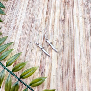 May include: A pair of silver-colored dagger earrings with a hook closure. The earrings are displayed on a light brown wooden surface, with green leaves in the background.