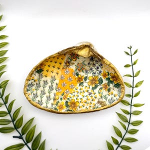Peut inclure: Un coquillage décoratif avec un bord doré. L'intérieur présente un motif floral avec des fleurs jaunes, blanches et lavande, et des abeilles. Le coquillage est posé sur un fond blanc avec du feuillage vert.