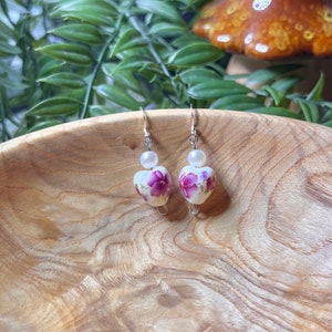 May include: A pair of heart-shaped earrings with a white background and purple floral design. Each earring features a white pearl bead and a silver hook.