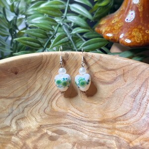 May include: A pair of white heart-shaped earrings with green floral designs and a white pearl bead on each earring.