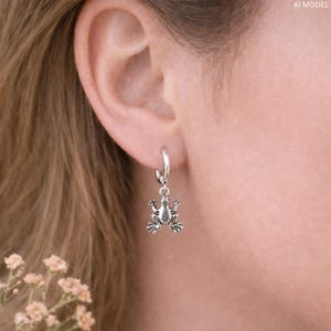 May include: A silver hoop earring with a small frog charm. The frog charm is detailed with visible legs, eyes, and skin texture. The earring is worn on a person's ear, with the focus on the jewelry.