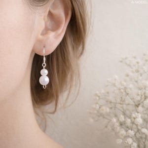 May include: A pair of elegant drop earrings featuring two white, luminous pearls suspended from a silver hook. The earrings are shown against a soft, neutral background, highlighting their classic design and delicate appeal. The pearls are approximately 0.5 inches in diameter.