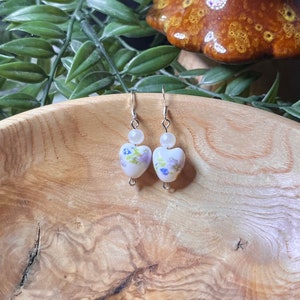 May include: A pair of silver dangle earrings with white heart-shaped beads featuring a floral pattern in shades of purple and green. The beads are accented with a single white pearl bead above each heart.