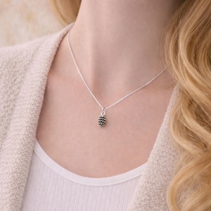 May include: A silver necklace with a small, textured pinecone pendant. The pendant is dark grey and suspended from a fine silver chain. The necklace is shown on a person wearing a white top and a light beige cardigan.