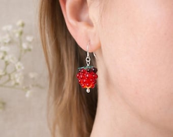 May include: A single earring featuring a red raspberry design. The earring has a silver hook and a cluster of red and dark red beads, with a green stem. The earring is worn on a person's ear.