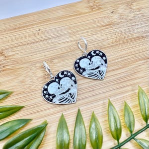 Puede incluir: Un par de pendientes en forma de corazón con un diseño de esqueleto. Los pendientes presentan un fondo negro con una pareja de esqueletos blancos abrazándose, rodeados de estrellas y lunas. Los pendientes son plateados con cierre de palanca.