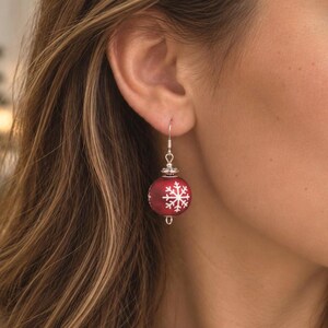 May include: A dangling earring featuring a spherical red bead decorated with a white snowflake pattern. The bead is suspended from a silver-toned metal hook and accented with a small silver-toned decorative cap at the top of the sphere.