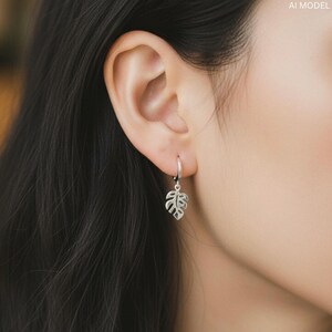 May include: A silver hoop earring with a dangling leaf charm. The leaf charm is a detailed cutout of a Monstera plant leaf. The earring is shown on a person's ear, with dark hair in the background.